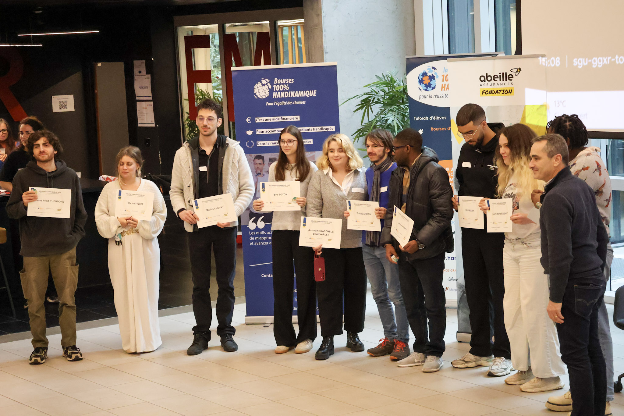 La photo montre une remise de prix ou de bourses d'études réunissant un groupe de personnes posant fièrement en ligne dans une salle lumineuse.