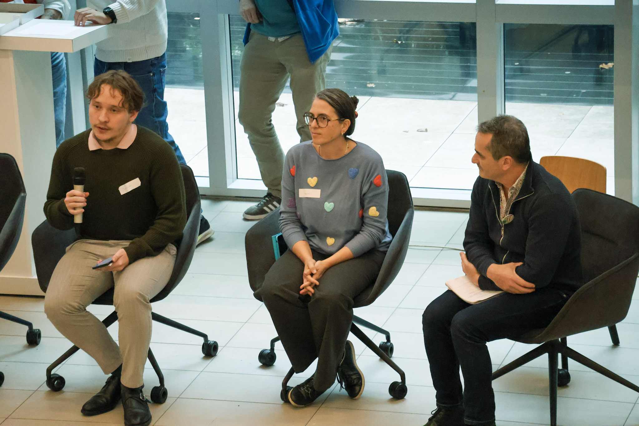 La photo montre une table ronde ou un panel de discussion lors d'une conférence en intérieur. Trois intervenants sont assis côte à côte face au public, sur des fauteuils de bureau noirs pivotants à roulettes, disposés sur un sol carrelé clair. Derrière eux se trouve une grande baie vitrée laissant entrer la lumière naturelle.
