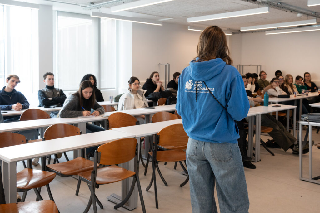 La scène se déroule dans une salle de cours ou de formation moderne, vaste et très éclairée par de grandes fenêtres sur la gauche. Elle montre une intervenante debout qui s'adresse à un groupe d'étudiants assis en rangées.