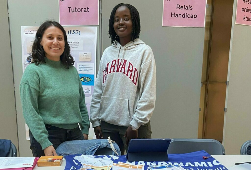 La photo montre deux jeunes femmes debout côte à côte, souriantes et accueillantes, derrière une table d'information lors d'un forum ou d'une permanence étudiante en intérieur.
