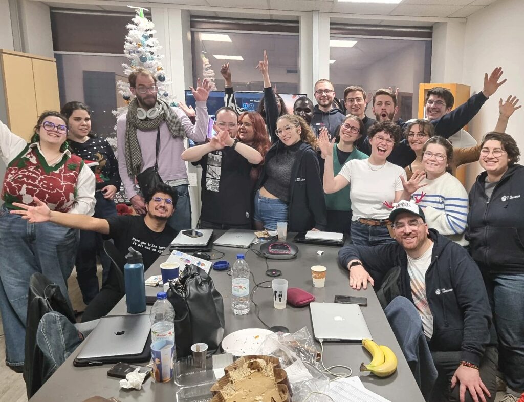 La photo capture une photo de groupe joyeuse et très animée, réunissant une quinzaine de jeunes adultes dans une salle de réunion à l'ambiance chaleureuse de fin d'année ou de fête de Noël.