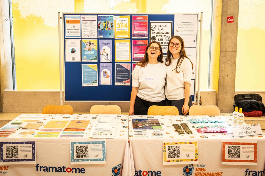 La scène montre deux jeunes femmes souriantes tenant un stand de prévention et de sensibilisation en intérieur, dans un espace bien éclairé par une grande baie vitrée en arrière-plan.