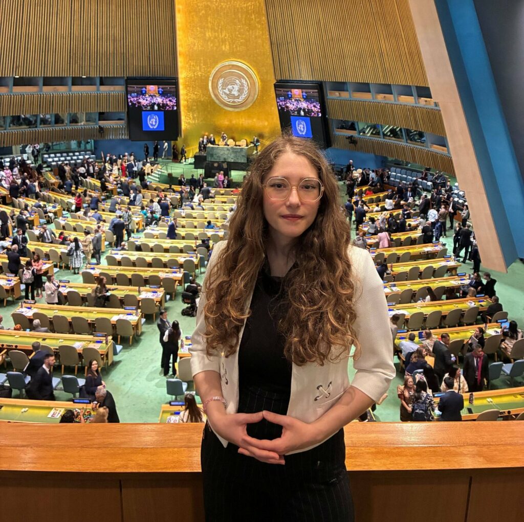 La photographie montre une jeune femme posant fièrement au premier plan d'un lieu hautement symbolique et prestigieux : la célèbre salle de l'Assemblée générale des Nations Unies (ONU) à New York.
