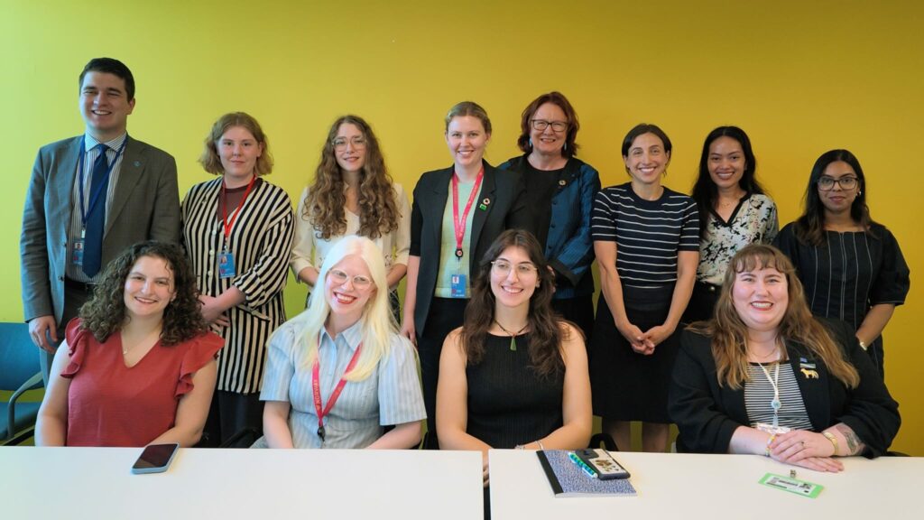 La scène montre une photo de groupe soignée et institutionnelle réunissant onze personnes dans une salle au fond jaune vif très lumineux. Le groupe, composé d'un homme et de dix femmes, est disposé sur deux rangées : quatre personnes assises au premier plan derrière une table blanche, et sept personnes debout au second plan.
