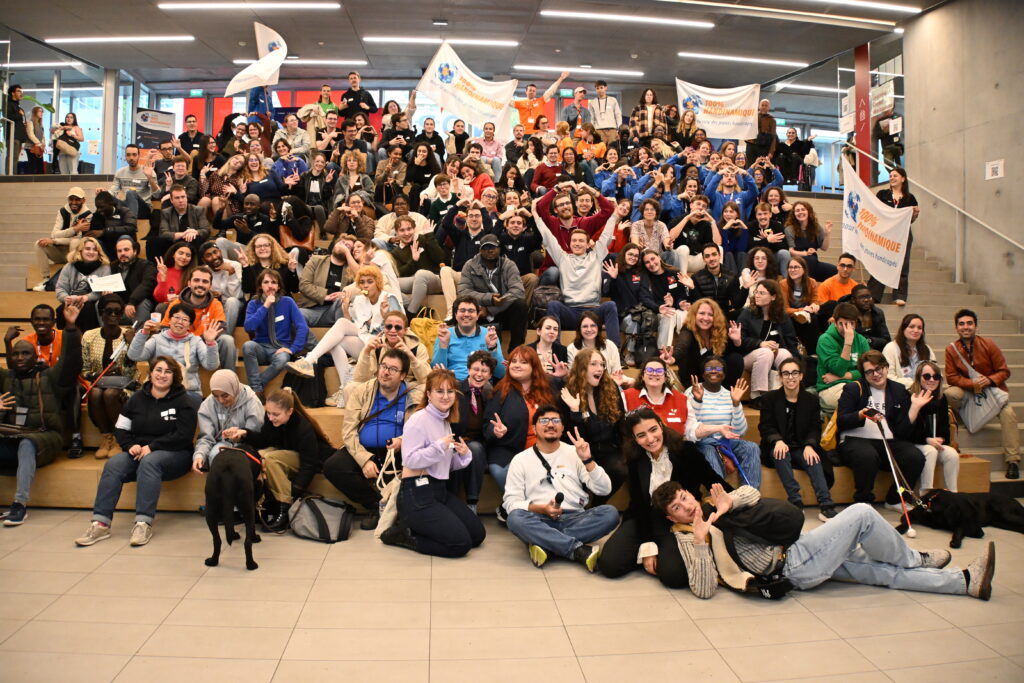 Il s'agit d'une très grande photo de groupe en intérieur, extrêmement vivante et festive, réunissant près d'une centaine de personnes sur de larges gradins en bois clair dans le hall d'un bâtiment moderne. L'atmosphère générale respire l'énergie, l'inclusion et la convivialité d'un grand rassemblement associatif.