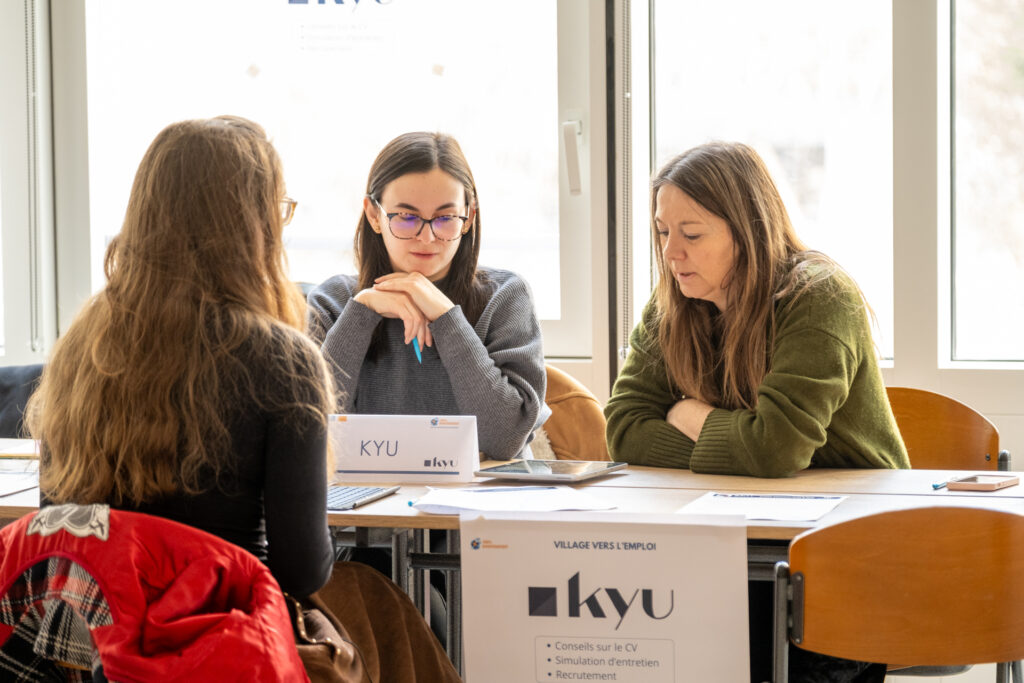 La scène illustre un entretien professionnel ou un atelier de coaching pour l'emploi autour d'une table, dans une salle lumineuse baignée par la lumière du jour provenant d'une grande fenêtre en arrière-plan.