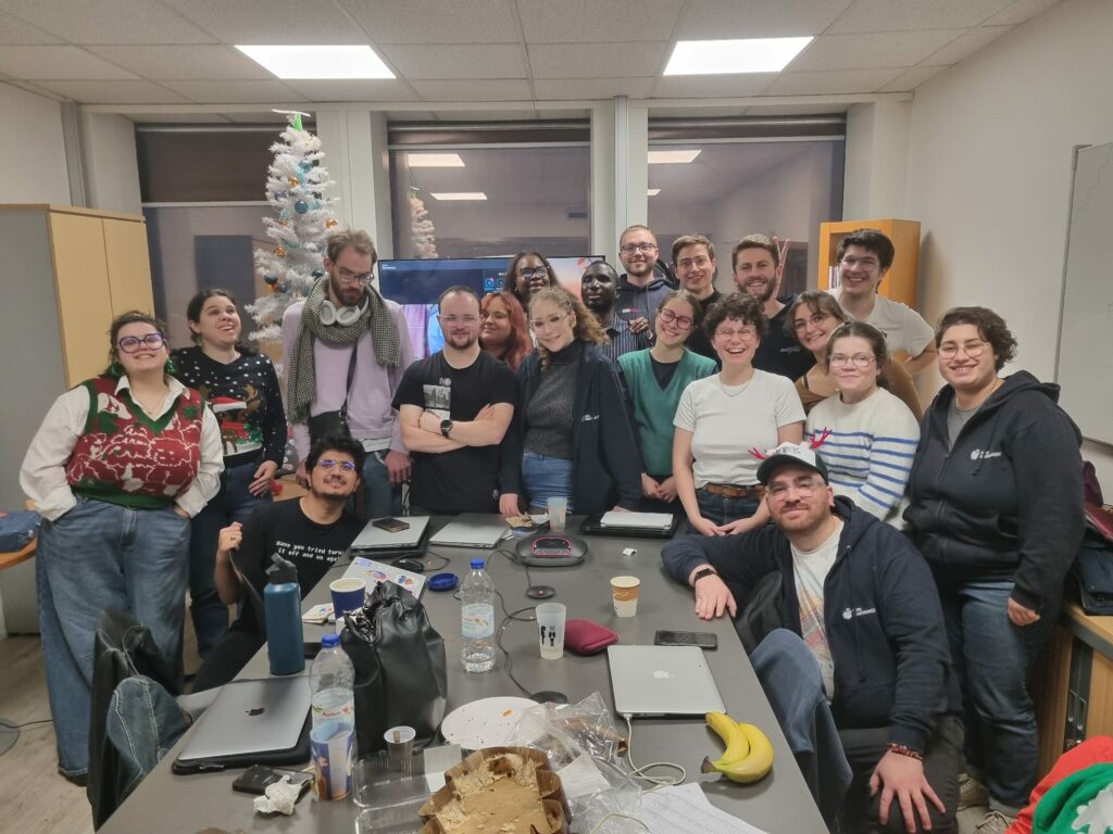 La photographie capture une photo de groupe chaleureuse et complice réunissant une quinzaine de personnes dans une salle de réunion décorée pour la période des fêtes de fin d'année. C'est une autre prise de vue de l'équipe réunie autour de la grande table de conférence.