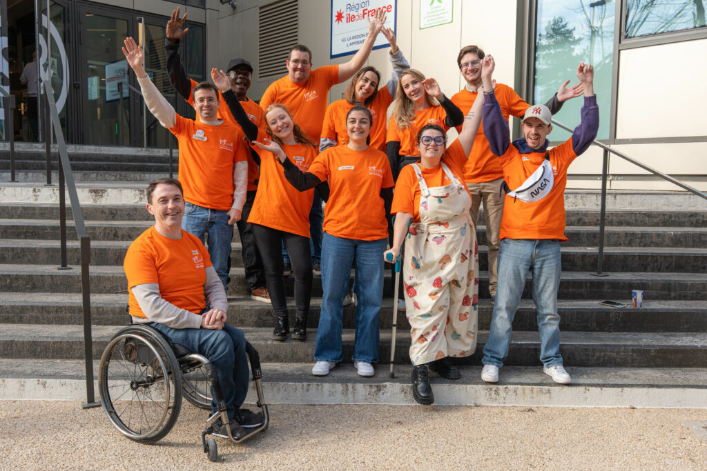 La scène est une photo de groupe en extérieur, prise en plein jour sur les marches d'un bâtiment moderne, rassemblant une dizaine de personnes vêtues de t-shirts orange vif et affichant une ambiance très chaleureuse, dynamique et solidaire.