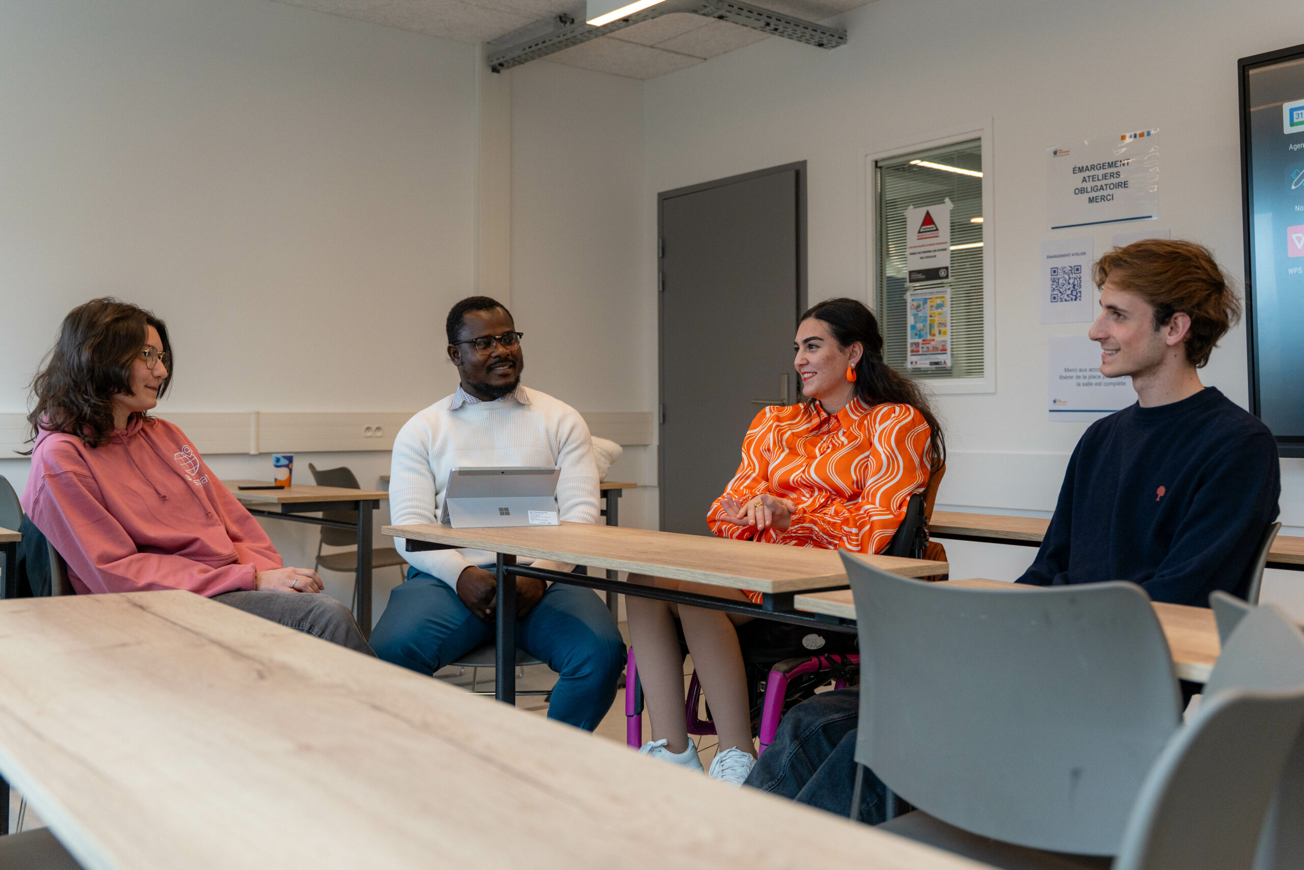 La scène se déroule en intérieur, dans une salle d'atelier ou de formation moderne et lumineuse. Quatre jeunes adultes sont réunis autour de tables en bois clair disposées pour un échange convivial en petit groupe, rappelant une séance de mentorat ou de travail d'équipe.