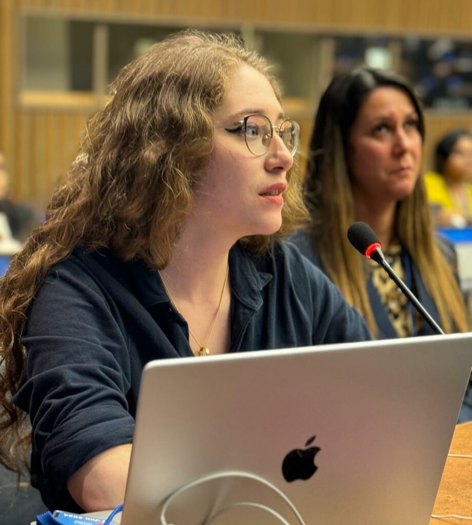 La scène montre une jeune femme prenant la parole lors d'une assemblée officielle, d'une conférence internationale ou d'une séance parlementaire.