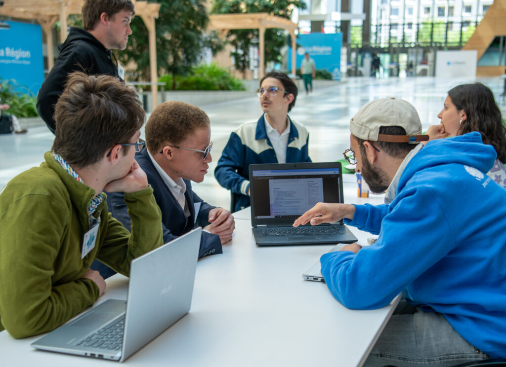 La scène montre un groupe de jeunes adultes réunis autour d'une table blanche lors d'une séance de travail collaboratif ou d'un atelier étudiant. Ils se trouvent dans le grand hall moderne et très lumineux d'un bâtiment public ou d'une institution régionale.