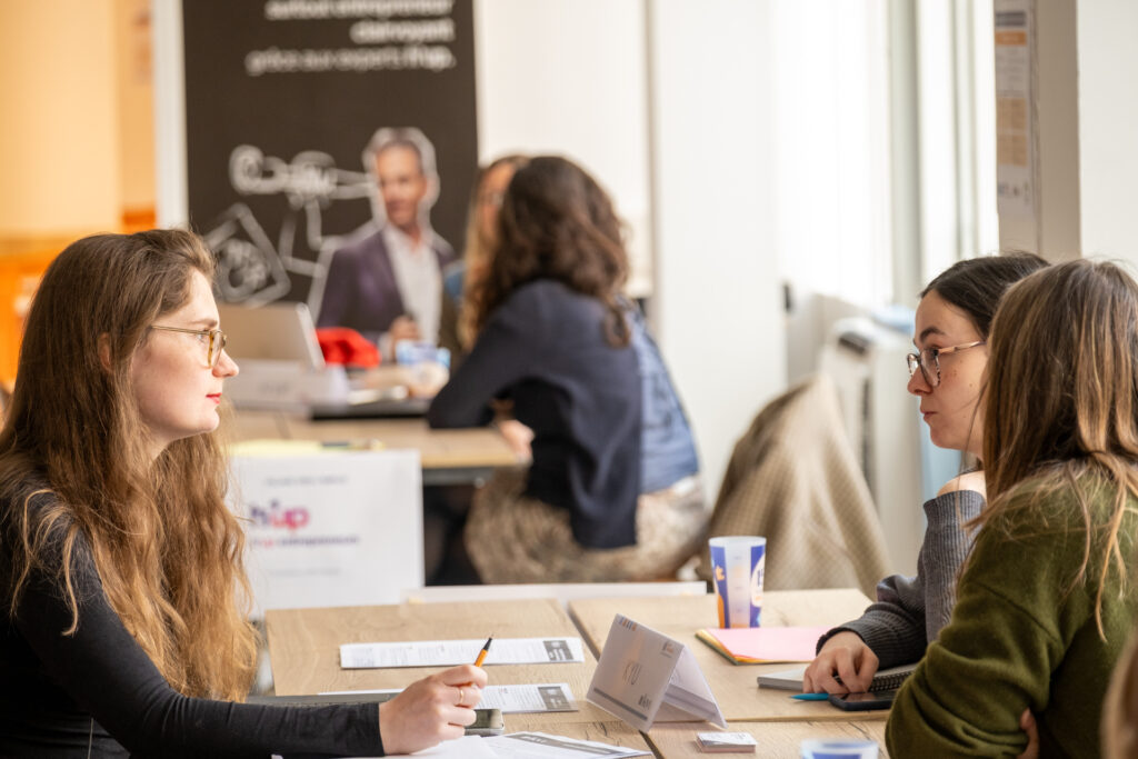 La scène montre un forum de rencontre professionnelle ou un atelier d'accompagnement pour l'emploi en intérieur, éclairé par la lumière naturelle d'une grande fenêtre située sur la droite. Plusieurs entretiens individuels s'y déroulent en même temps dans une ambiance sérieuse et attentive.