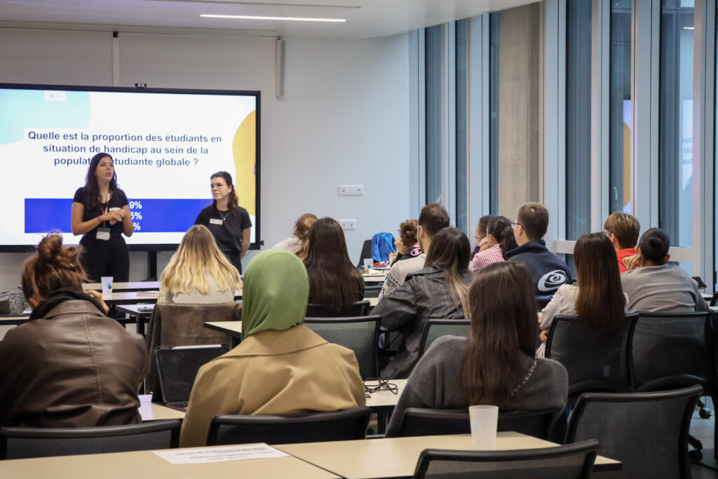 La scène se déroule en intérieur, dans une salle de conférence ou de formation moderne et très lumineuse. Elle montre une présentation interactive devant une assemblée d'étudiants ou de participants attentifs, assis de dos par rapport à l'objectif.