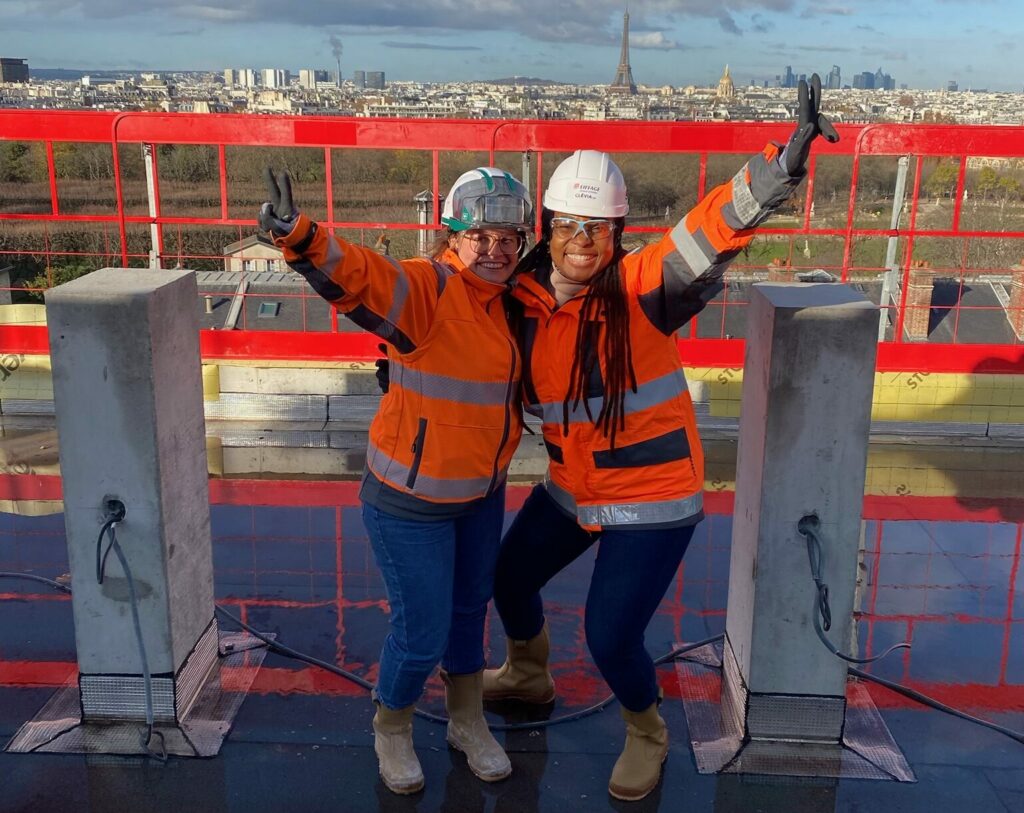 La scène est une photographie en plein air prise sur le toit-terrasse d'un chantier en hauteur, avec une vue panoramique impressionnante sur les toits de Paris. Deux femmes professionnelles du bâtiment ou en visite de chantier posent côte à côte, pleines d'énergie et de joie.