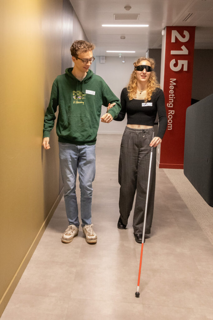 La scène se déroule en intérieur, le long d'un couloir de bureaux moderne et bien éclairé. Elle montre un exercice de guidage ou un atelier de sensibilisation à la déficience visuelle entre deux jeunes adultes marchant côte à côte vers l'objectif.