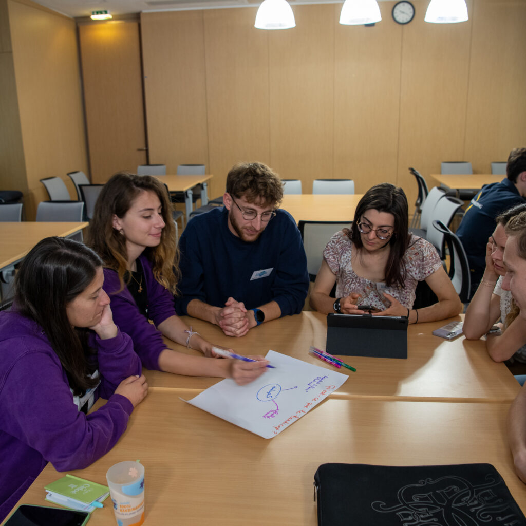 La scène montre un groupe d'étudiants ou de participants réunis autour d'une table en bois clair pour un atelier de réflexion collective ou un travail de groupe en sous-commission, dans une salle lumineuse aux murs lambrissés de bois clair.