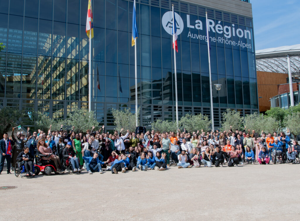 La scène est une immense photo de groupe prise en extérieur, par une belle journée ensoleillée, sur une vaste esplanade devant un grand bâtiment institutionnel moderne en verre. Elle rassemble plus d'une centaine de participants, d'étudiants, d'accompagnateurs et de bénévoles célébrant ensemble un grand événement associatif dans un élan de joie et de convivialité.