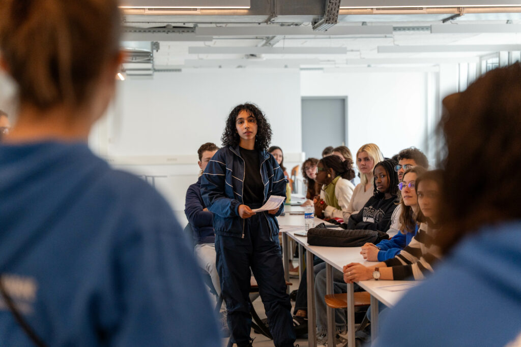 La scène se déroule dans une salle de formation ou de cours moderne et lumineuse. Elle montre une prise de parole interactive au milieu d'un groupe d'étudiants assis à des tables.