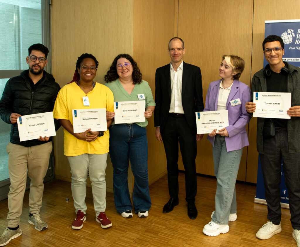 La photographie capture une cérémonie officielle de remise de bourses ou de prix en intérieur, réunissant six personnes alignées côte à côte et souriantes sur un plancher en parquet de bois clair. Cinq étudiants lauréats tiennent fièrement un certificat ou diplôme blanc rectangulaire aux côtés d'un représentant en costume.