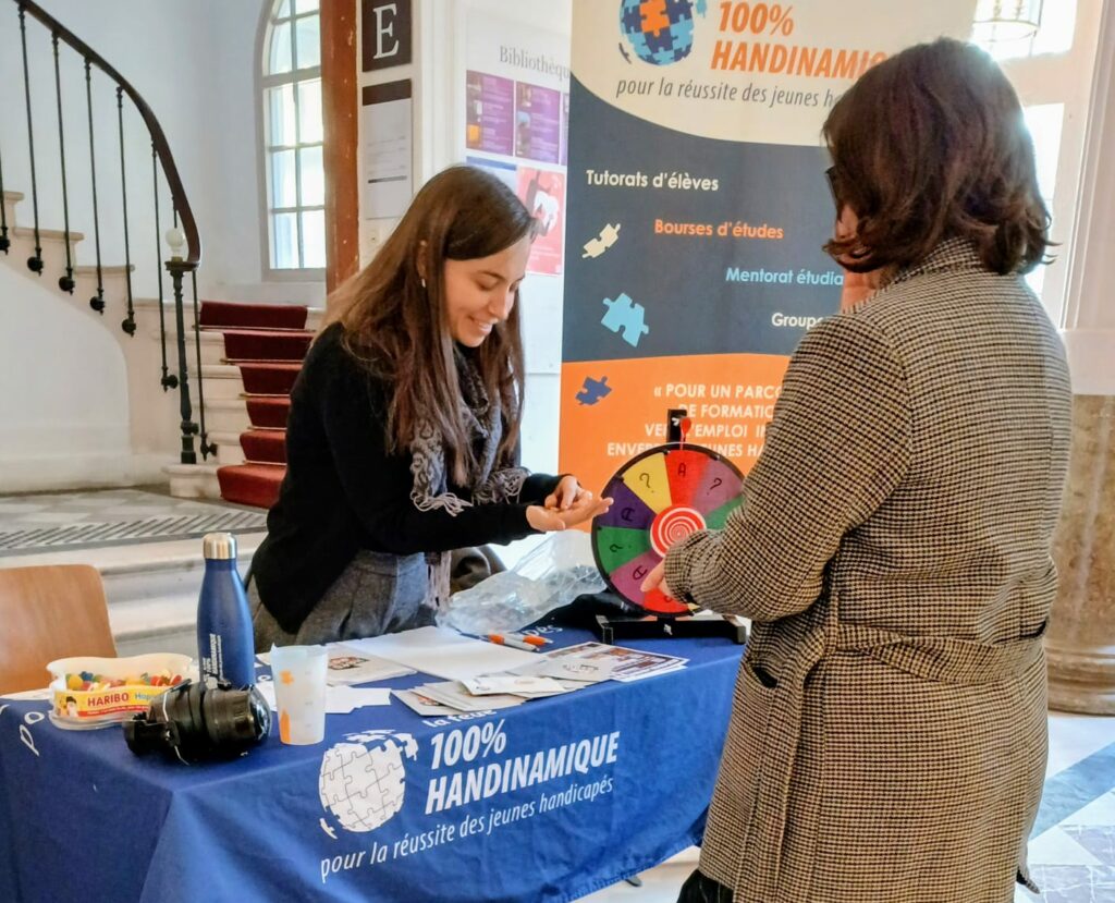 Deux femmes se parlent autour d'un stand associatif dans le hall lumineux d'un grand bâtiment. La scène se déroule dans le hall lumineux d'un bâtiment ancien ou d'une institution. À l'arrière-plan sur la gauche, un escalier monumental en pierre tourne doucement vers l'étage, bordé d'une rampe en fer forgé sombre et recouvert d'un tapis rouge. De hautes fenêtres laissent entrer une lumière naturelle claire.